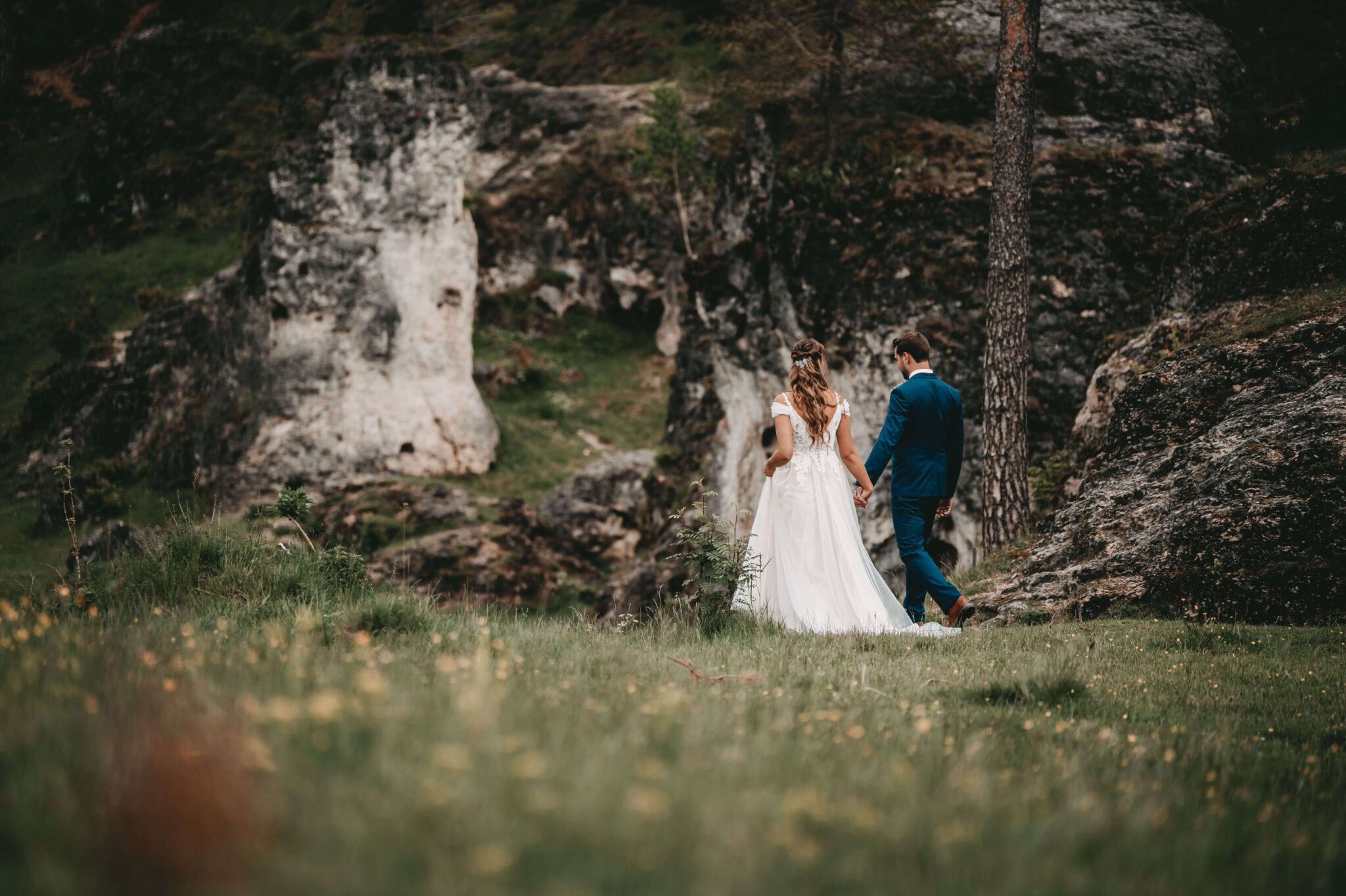 Ein Brautpaar spaziert Hand in Hand durch eine malerische Landschaft mit Felsen im Hintergrund. Die Braut trägt ein weißes Kleid und der Bräutigam einen blauen Anzug. Foto aufgenommen von Patrick Bertsche, Hochzeitsfotograf Mannheim.