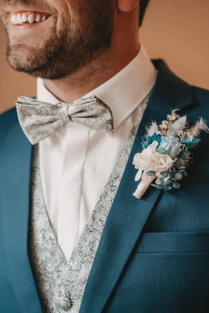 Eine Nahaufnahme des Bräutigams in einem eleganten blauen Anzug mit Fliege und Boutonnière, die seine Vorfreude auf den besonderen Moment zeigt.