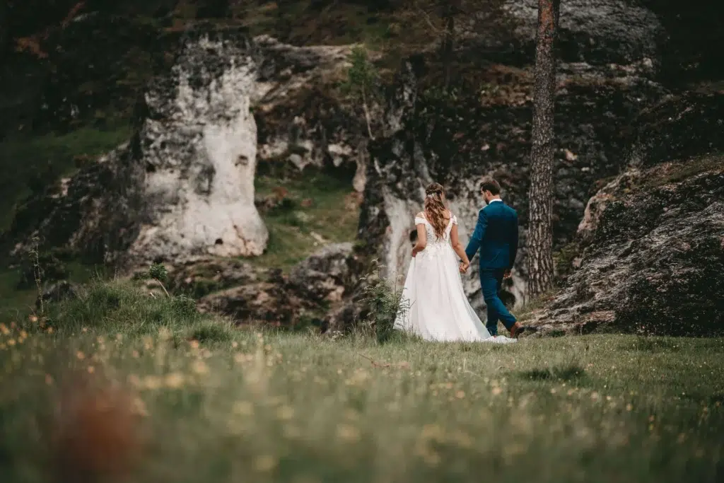 Ein Brautpaar spaziert Hand in Hand durch eine malerische Landschaft, umgeben von Felsen und Wiesenblumen.