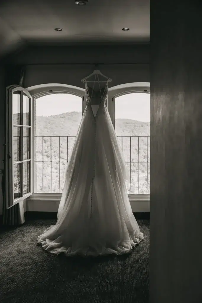 Ein elegantes Hochzeitskleid hängt an einem Fensterrahmen, durch das die sanften Hügel der Landschaft sichtbar sind. Das Kleid ist perfekt drapiert und wartet auf den großen Moment der Braut, während das offene Fenster eine ruhige, romantische Atmosphäre schafft.