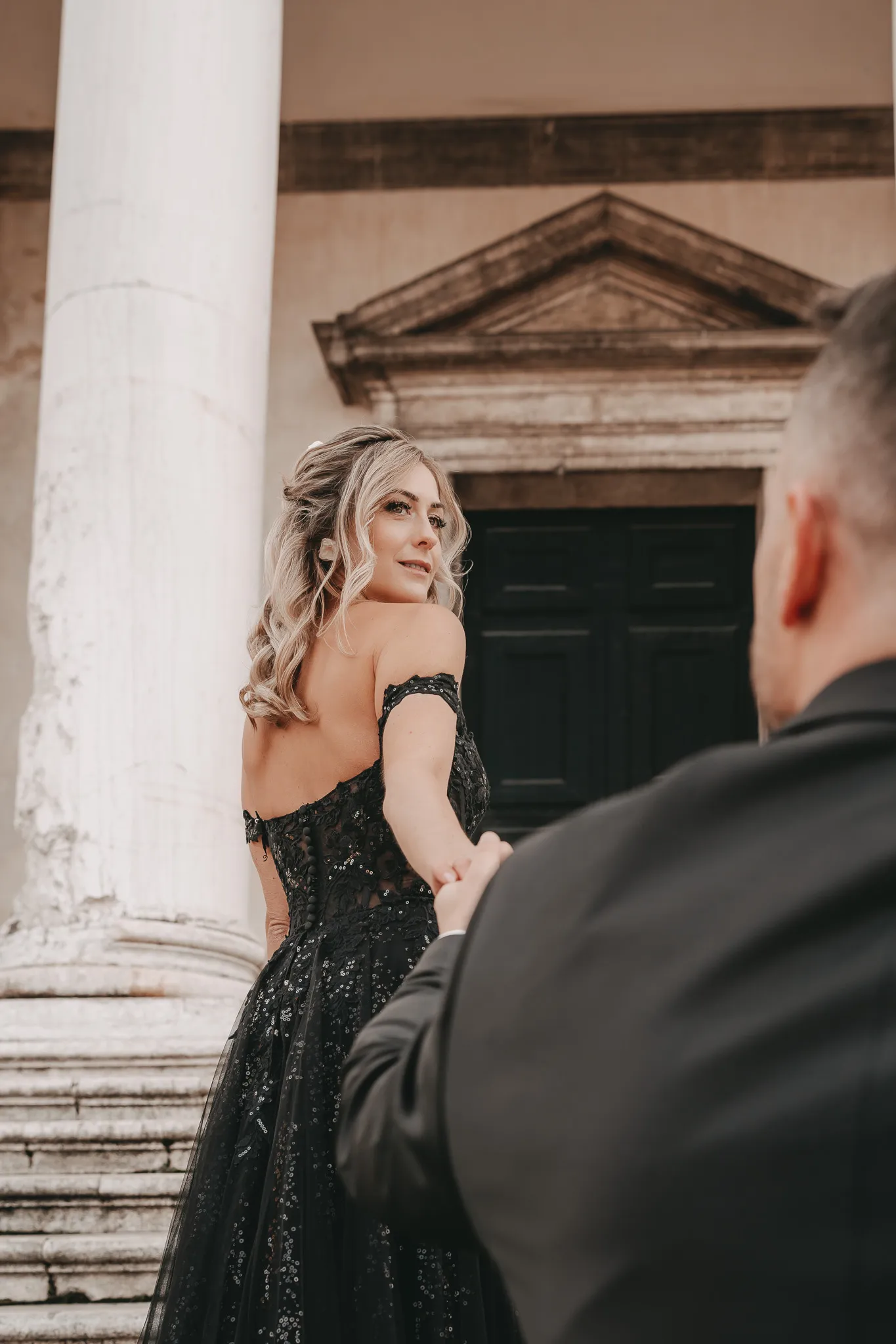 Die Braut in einem schwarzen Hochzeitskleid blickt liebevoll über die Schulter, während sie die Hand des Bräutigams hält – Romantik in Venedig.